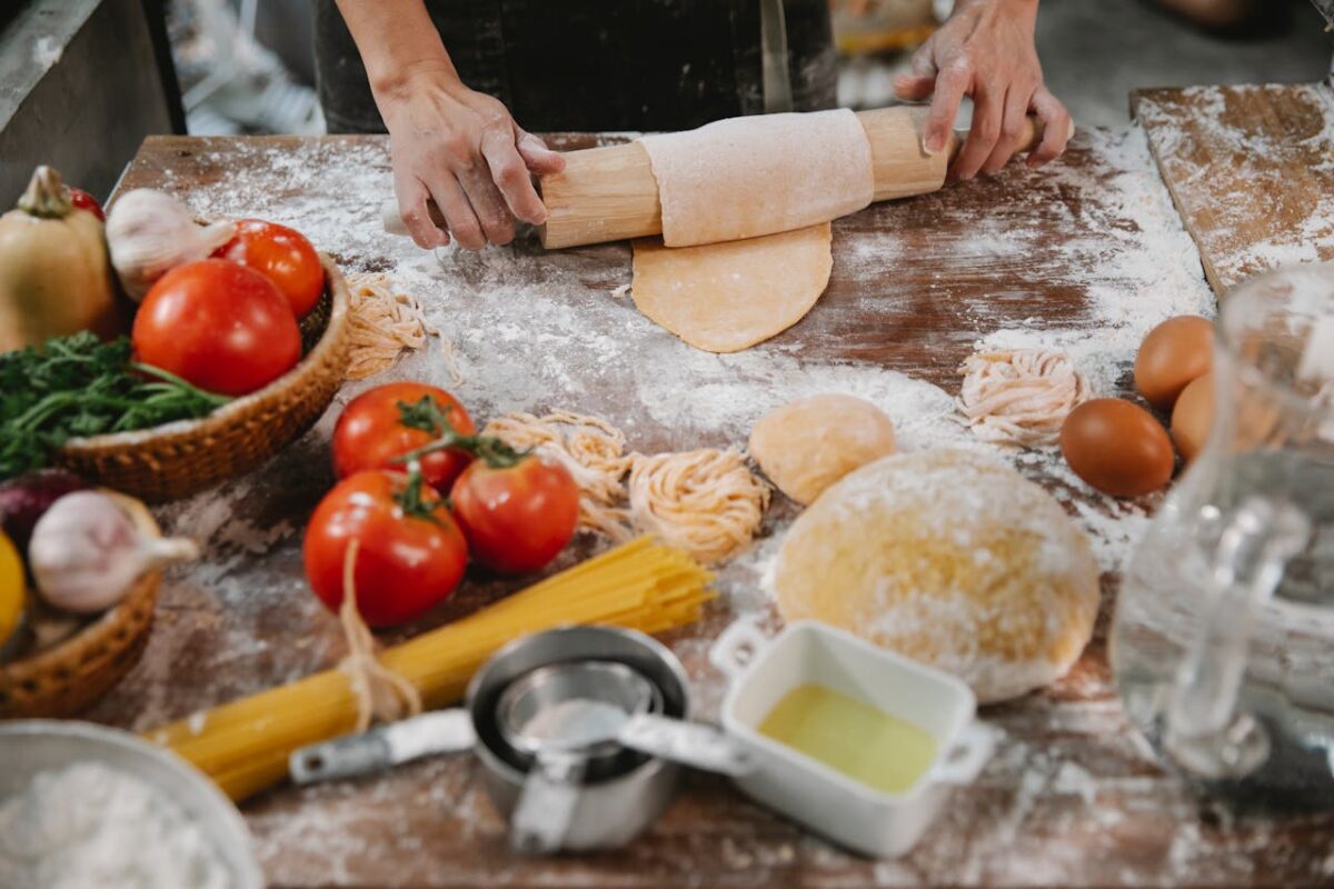 Crop anonymous female cooking homemade pasta on table covered with flour among vegetables