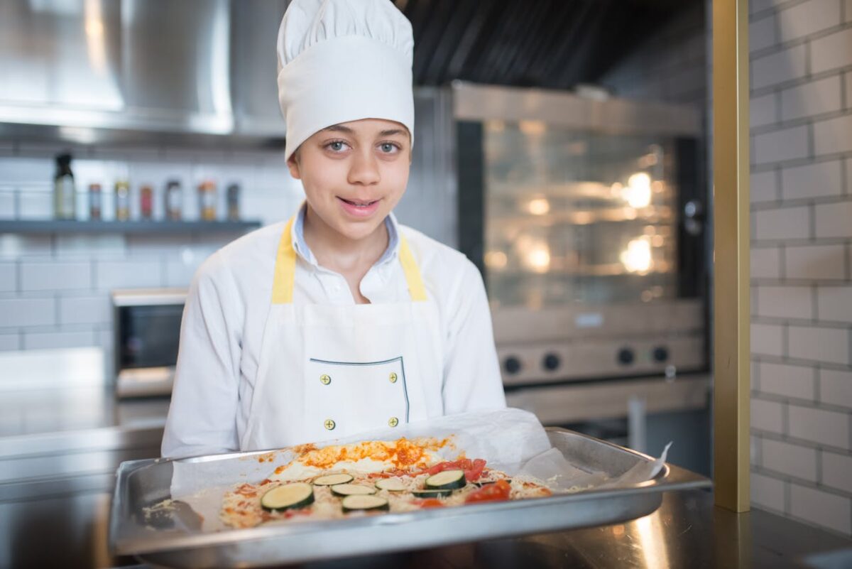 A young boy wearing a chef outfit prepares a pizza in a professional kitchen setting.